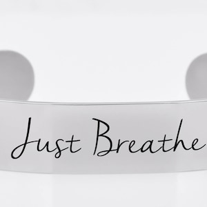 May include: A silver metal cuff bracelet with the words "Just Breathe" engraved on the front.