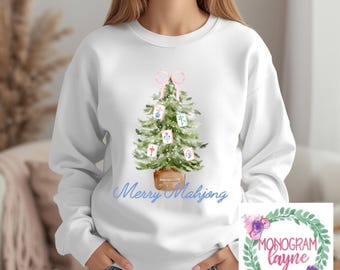 May include: White sweatshirt featuring a watercolor Christmas tree decorated with pink bow and Mahjong tiles. The phrase "Merry Mahjong" is written in blue script. A floral wreath with the text "Monogram Layne" is in the lower right corner.