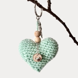 Puede incluir: Un llavero de corazón tejido a crochet de color verde claro con cuentas de madera y un botón. El llavero está unido a un llavero de plata.