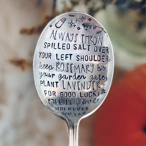 Puede incluir: Una cuchara plateada con texto estampado. El texto dice: "ALWAYS THROW SPILLED SALT OVER YOUR LEFT SHOULDER, keep ROSEMARY by your garden gate, PLANT LAVENDER FOR GOOD LUCK, & FALL IN LOVE WHENEVER YOU CAN." Una calabaza naranja y flores secas están en el fondo.