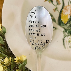May include: A silver-toned metal spoon featuring the stamped text IN A WORLD WHERE YOU CAN BE ANYTHING BE A dolly, accented with small star and heart symbols. The spoon has a decorative handle and is positioned vertically over a floral plate.