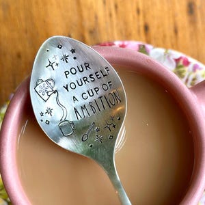 Puede incluir: Una cuchara de metal plateado con un dise&ntilde;o estampado que muestra una regadera, una taza peque&ntilde;a y el texto "POUR YOURSELF A CUP OF AMBITION" rodeado de estrellas y diamantes, descansando dentro de una taza de cer&aacute;mica rosa con l&iacute;quido marr&oacute;n claro.