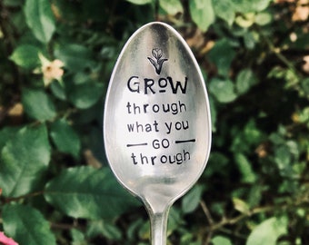 Puede incluir: Una cuchara de plata con las palabras "Grow through what you go through" grabadas en ella. La cuchara está delante de un fondo verde frondoso.