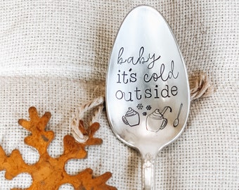 May include: A silver spoon with the text "baby it's cold outside" engraved on it. The spoon is decorated with a snowflake and two mugs of hot chocolate.
