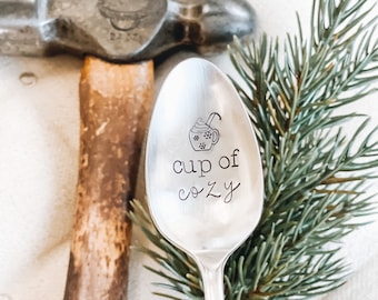 May include: A silver spoon with a design of a cup of hot cocoa and the text "cup of cozy" engraved on it. The spoon is sitting on a wooden surface with a green pine branch in the background.