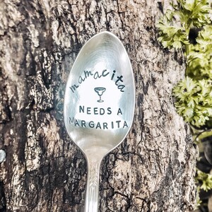 Puede incluir: Una cuchara de plata con el texto "mamacita needs a margarita" grabado en ella. La cuchara tiene un pequeño icono de vaso de margarita sobre el texto.