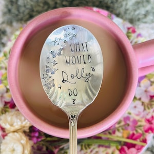 May include: A silver-toned metal spoon with a scalloped edge, featuring the stamped black text "WHAT WOULD Dolly DO ?" and small engraved butterfly and star icons on the bowl, resting inside a pink ceramic mug filled with a light brown liquid.