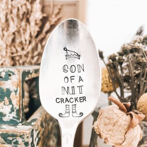 Peut inclure: Une cuillère en argent gravée avec le texte "Son of a Nut Cracker". La cuillère a un petit chapeau d'elfe et des chaussures d'elfe en haut et en bas du texte.