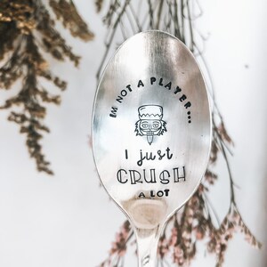 Peut inclure: Une cuillère en argent avec le texte estampé "I'M NOT A PLAYER... I JUST CRUSH A LOT." Un visage de dessin animé est estampé au-dessus des mots "I just". La cuillère est posée sur un fond flou de fleurs séchées.
