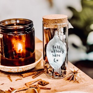 Op de afbeelding: Een glazen pot met een kurken deksel gevuld met gedroogde kruiden en een zilveren lepel met de tekst "fell asleep here" en een halve maan en een ster. De pot staat op een houten oppervlak met kaneelstokjes en een gedroogde bloem.