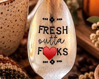 Puede incluir: Una cuchara de metal plateado con el texto negro estampado FRESH outta FKS y un icono de corazón rojo que sustituye a la letra U. El mango de la cuchara presenta un diseño floral en relieve en el cuello y la pala está centrada.