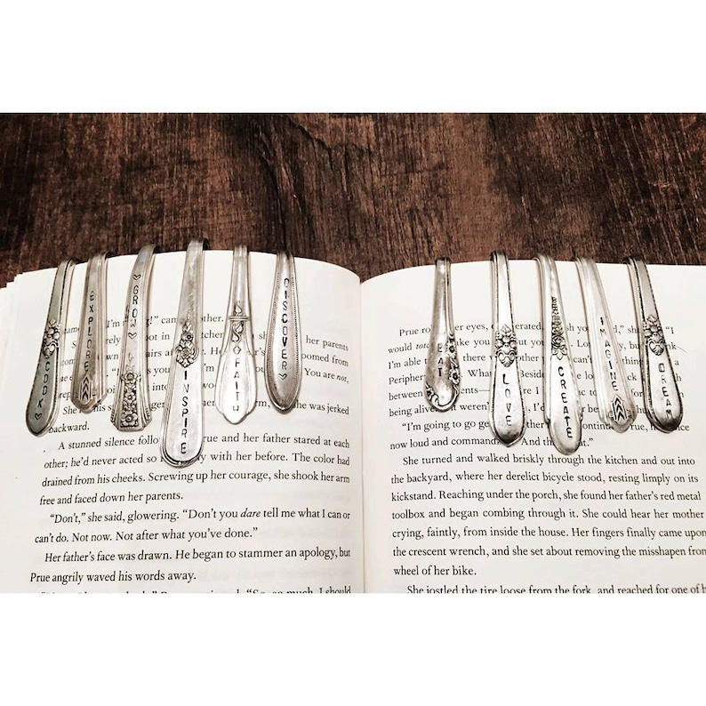 Pode incluir: Uma cole&ccedil;&atilde;o de colheres de prata com palavras como "Discover", "Love", "Create" e "Inspire" gravadas nelas. As colheres s&atilde;o usadas como marcadores de livros e s&atilde;o colocadas em um livro aberto.