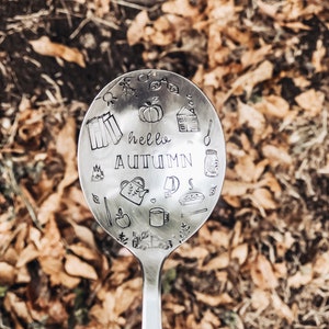 Puede incluir: Una cuchara de plata con un diseño de ilustraciones de otoño y el texto "hello autumn". La cuchara está sobre un lecho de hojas marrones.