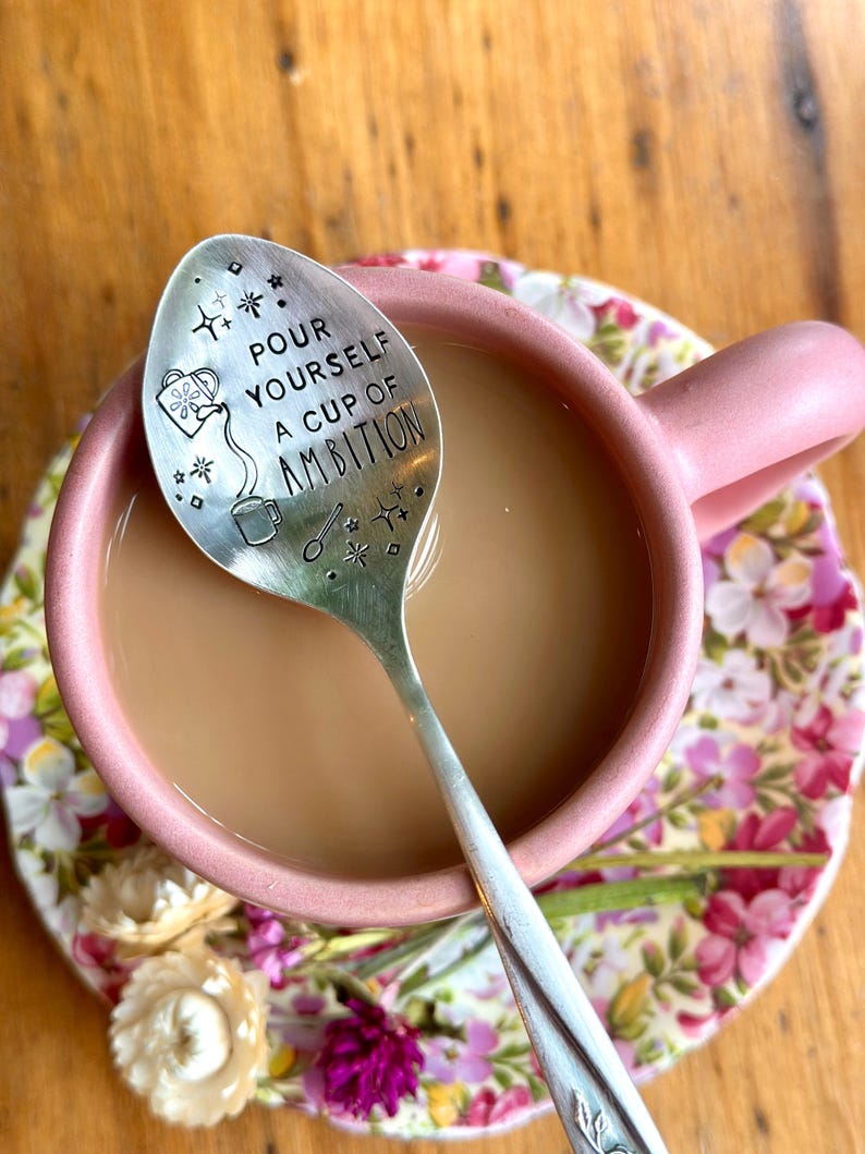 Puede incluir: Una cuchara de metal plateado con un dise&ntilde;o estampado que muestra una regadera, una taza peque&ntilde;a y el texto "POUR YOURSELF A CUP OF AMBITION" rodeado de estrellas y diamantes, descansando dentro de una taza de cer&aacute;mica rosa con l&iacute;quido marr&oacute;n claro.