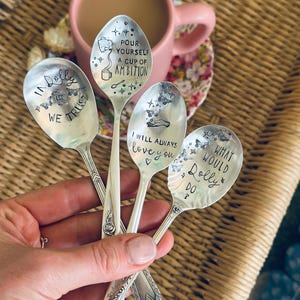 Puede incluir: Cuatro cucharas de metal plateado con textos y dise&ntilde;os estampados. Las cucharas presentan las frases: IN DOLLY WE TRUST, POUR YOURSELF A CUP OF AMBITION, I WILL ALWAYS LOVE YOU y WHAT WOULD DOLLY DO, decoradas con mariposas, estrellas y corazones.