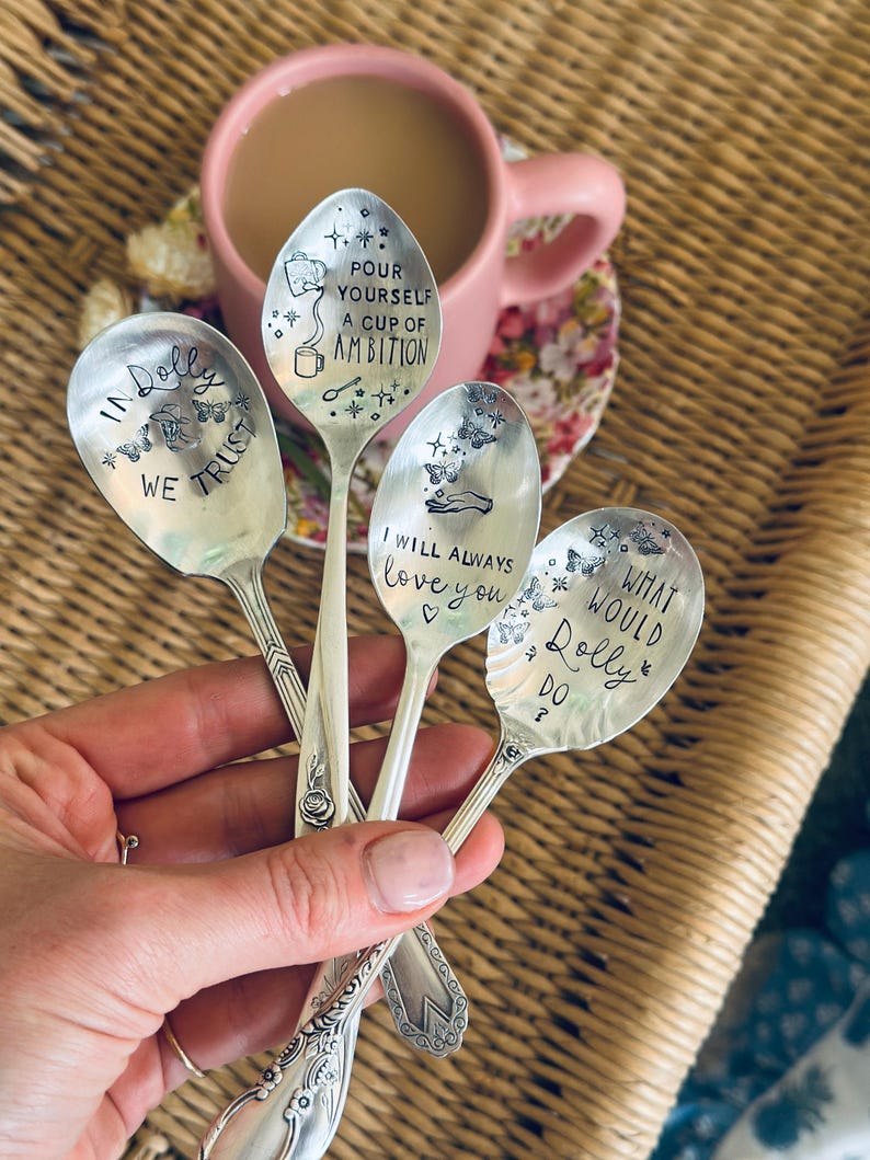 Puede incluir: Cuatro cucharas de metal plateado con textos y dise&ntilde;os estampados. Las cucharas presentan las frases: IN DOLLY WE TRUST, POUR YOURSELF A CUP OF AMBITION, I WILL ALWAYS LOVE YOU y WHAT WOULD DOLLY DO, decoradas con mariposas, estrellas y corazones.