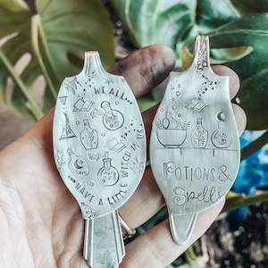 May include: Two silver spoons with engraved designs. One spoon has the text "We all... have a little witch in us." and images of potion bottles, books, and moons. The other spoon has the text "Potions & Spells" and images of potion bottles, a cauldron, and a book.