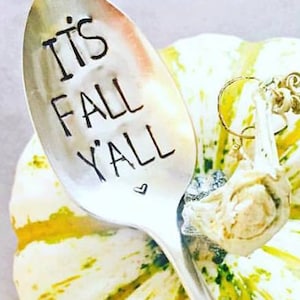 Op de afbeelding: Een zilveren lepel met de tekst "It's Fall Y'all" en een hartsymbool. De lepel ligt op een decoratieve kalebas met groene en gele patronen. Er hangt een kleine bedel aan de lepel.