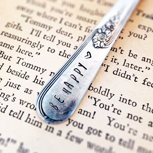 Pode incluir: Uma colher de prata com as palavras "Be Happy" gravadas nela, descansando sobre uma p&aacute;gina de um livro. A colher serve como marcador de p&aacute;gina.