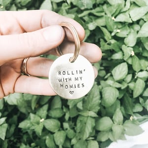 Puede incluir: Un llavero dorado con una etiqueta redonda que dice "ROLLIN' WITH MY HOMIES" con un pequeño símbolo de corazón.