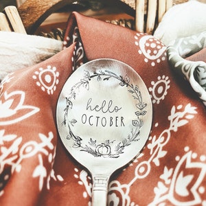 Op de afbeelding: Een zilveren lepel met een bloemmotief en de woorden "hello october" erop gegraveerd. De lepel ligt op een rood-witte gepatenteerde stof.