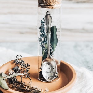 Op de afbeelding: Een glazen buis met een kurken stop bevat een zilveren lepel met de inscriptie "The magic is in you, babe". De buis staat op een terracotta schotel met gedroogde lavendel en eucalyptus.