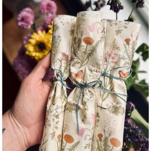 Puede incluir: Tres rollos cil&iacute;ndricos de papel color crema con un estampado bot&aacute;nico de dientes de le&oacute;n naranjas, flores silvestres rosas y mariposas marrones. Cada rollo est&aacute; atado con un cordel fino de color azul verdoso oscuro.