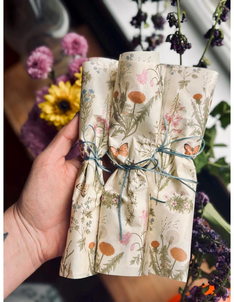 Puede incluir: Tres rollos cil&iacute;ndricos de papel color crema con un estampado bot&aacute;nico de dientes de le&oacute;n naranjas, flores silvestres rosas y mariposas marrones. Cada rollo est&aacute; atado con un cordel fino de color azul verdoso oscuro.