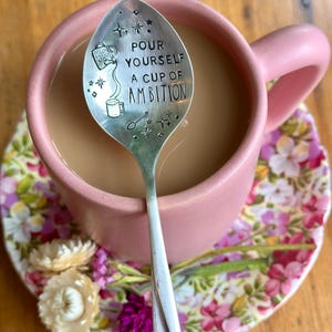 Puede incluir: Una cuchara de metal plateado con el texto estampado "POUR YOURSELF A CUP OF AMBITION" dentro de una taza rosa mate llena de caf&eacute;. La cuchara presenta ilustraciones grabadas de una regadera, una taza peque&ntilde;a y varias formas de estrellas.