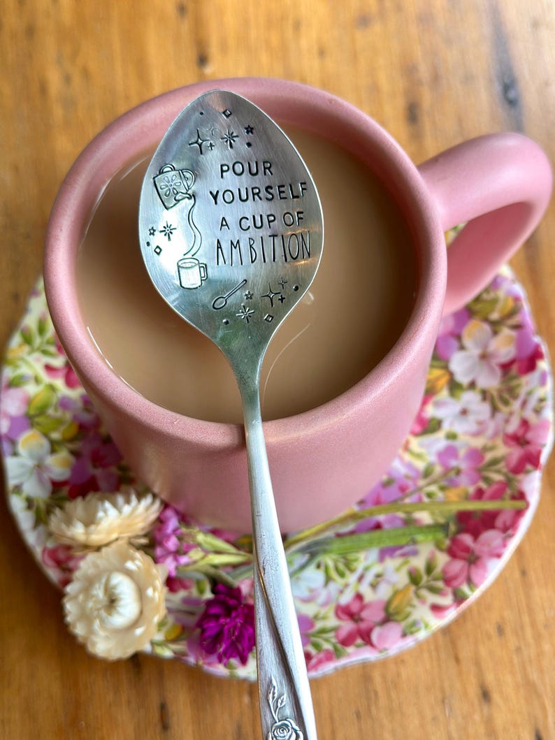 Puede incluir: Una cuchara de metal plateado con el texto estampado "POUR YOURSELF A CUP OF AMBITION" dentro de una taza rosa mate llena de caf&eacute;. La cuchara presenta ilustraciones grabadas de una regadera, una taza peque&ntilde;a y varias formas de estrellas.