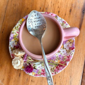 Puede incluir: Una cuchara de metal plateado con un motivo de rosa en el mango y el texto "POUR YOURSELF A CUP OF AMBITION" grabado en la cazoleta, descansando en una taza de cer&aacute;mica rosa con caf&eacute;, sobre un plato floral con peque&ntilde;as flores secas.