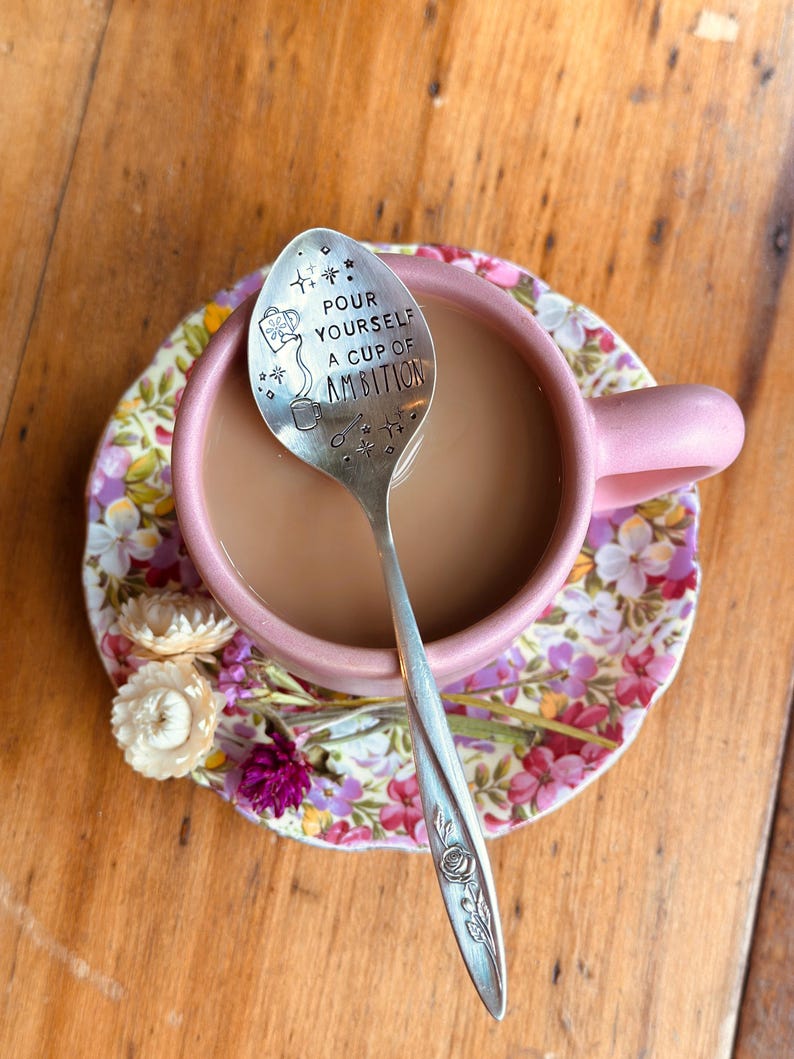 Puede incluir: Una cuchara de metal plateado con un motivo de rosa en el mango y el texto "POUR YOURSELF A CUP OF AMBITION" grabado en la cazoleta, descansando en una taza de cer&aacute;mica rosa con caf&eacute;, sobre un plato floral con peque&ntilde;as flores secas.