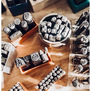 Puede incluir: Una colecci&oacute;n de herramientas de estampado de metal dispuestas en varios recipientes. El conjunto incluye numerosos punzones de acero con diversos dise&ntilde;os grabados como un oso, una abeja y patrones geom&eacute;tricos, junto con caracteres alfanum&eacute;ricos.