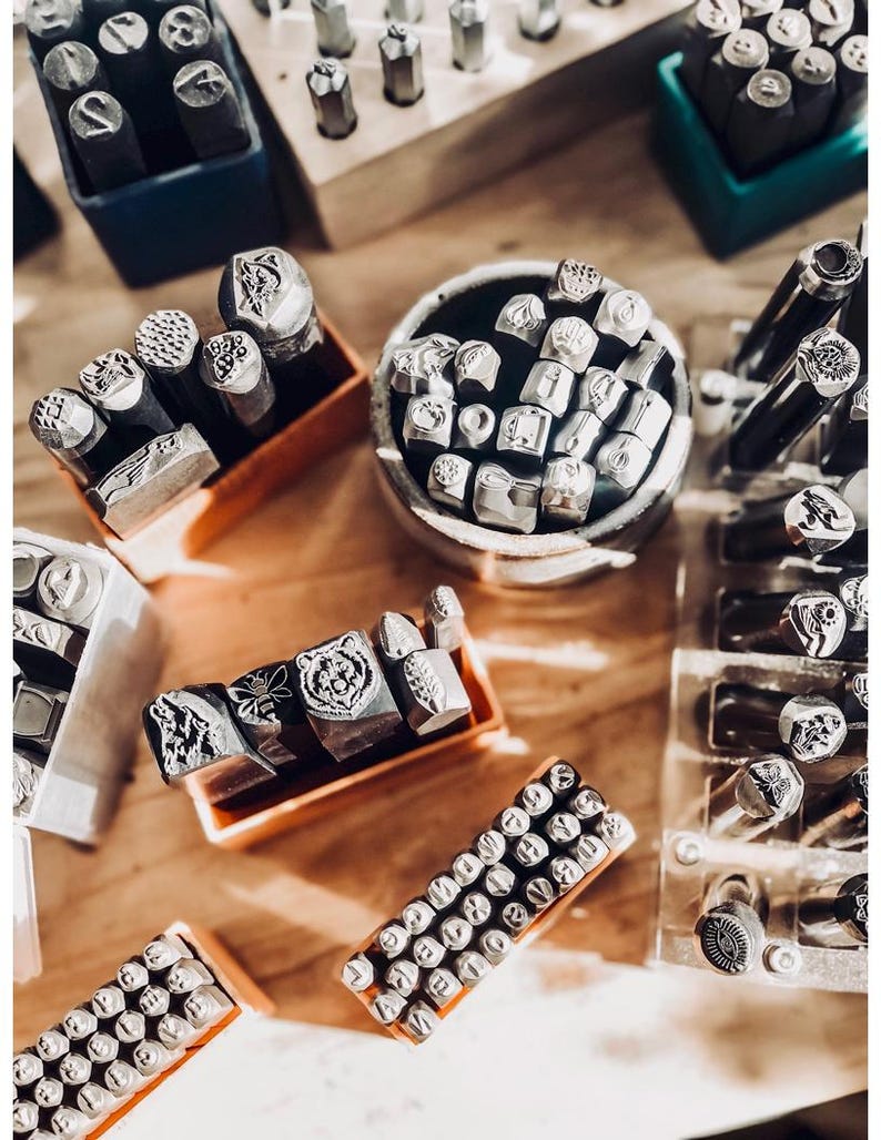 Puede incluir: Una colecci&oacute;n de herramientas de estampado de metal dispuestas en varios recipientes. El conjunto incluye numerosos punzones de acero con diversos dise&ntilde;os grabados como un oso, una abeja y patrones geom&eacute;tricos, junto con caracteres alfanum&eacute;ricos.