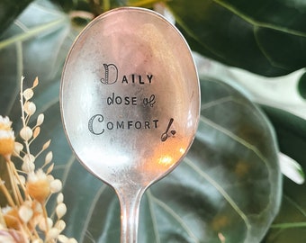 Puede incluir: Una cuchara de plata con las palabras "DAILY dose of COMFORT" grabadas en el cuenco. La cuchara está rodeada de follaje verde.