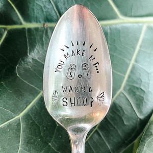 Op de afbeelding: Een zilveren lepel met de gestempelde tekst "YOU MAKE ME WANNA SHOOP". De lepel heeft illustraties van zout- en peperstrooiers en kleine bloemmotieven. De lepel staat tegen een groene, bladerrijke achtergrond.
