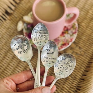 Puede incluir: Cuatro cucharas de metal plateado con texto e ilustraciones estampadas. Las cucharas presentan frases que incluyen "IN DOLLY WE TRUST", "POUR YOURSELF A CUP OF AMBITION", "I WILL ALWAYS love you" y "WHAT WOULD DOLLY DO?" decoradas con mariposas y estrellas.