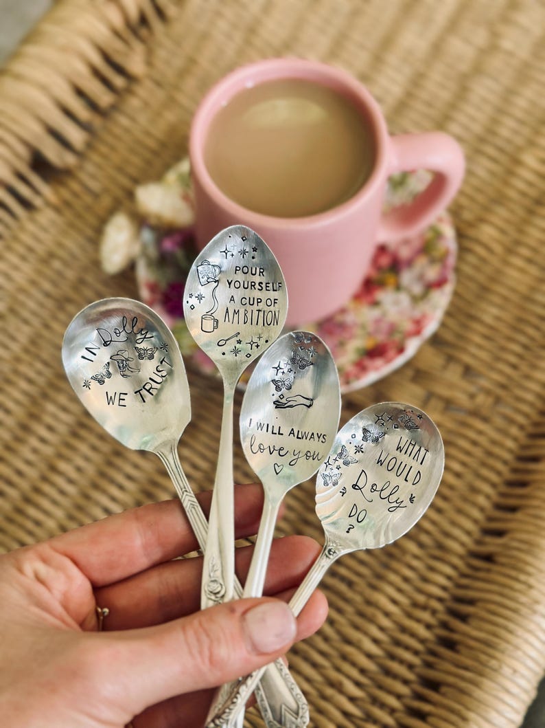 Puede incluir: Cuatro cucharas de metal plateado con texto e ilustraciones estampadas. Las cucharas presentan frases que incluyen "IN DOLLY WE TRUST", "POUR YOURSELF A CUP OF AMBITION", "I WILL ALWAYS love you" y "WHAT WOULD DOLLY DO?" decoradas con mariposas y estrellas.