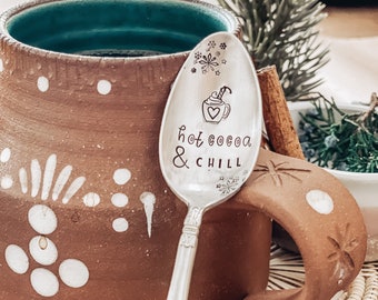 May include: A silver spoon with the words "hot cocoa & chill" engraved on it. The spoon is resting on the rim of a brown ceramic mug with white polka dots.