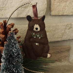 May include: A brown felt bear ornament holding a sprig of green holly with white berries. The bear is sitting on a white background.