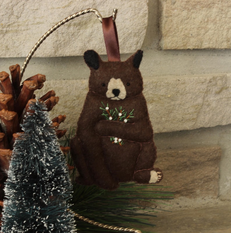 May include: A brown felt bear ornament holding a sprig of green holly with white berries. The bear is sitting on a white background.