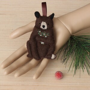 May include: A brown felt bear ornament holding a sprig of green holly with white berries. The ornament has a brown ribbon loop for hanging.
