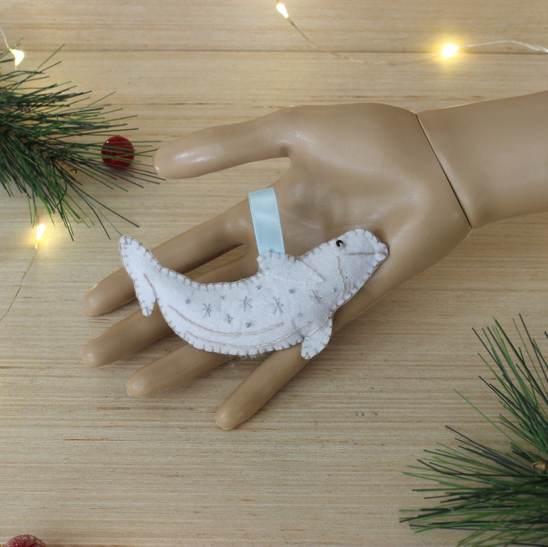 May include: White felt ornament shaped like a beluga whale with a blue ribbon. The whale has a white felt body with silver snowflake embroidery.