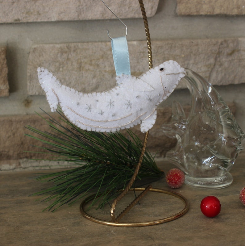 May include: White felt ornament shaped like a beluga whale with a blue ribbon for hanging. The whale has a white felt body with silver glitter snowflakes embroidered on it. The ornament is displayed on a gold metal stand.