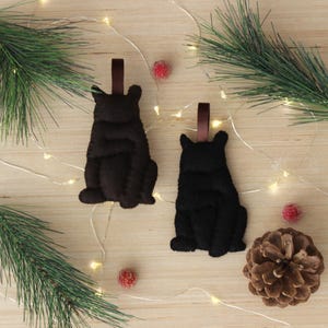 May include: Two felt bear ornaments, one brown and one black, hanging from brown ribbon. The bears are sitting with their paws tucked in front of them. The ornaments are on a wooden background with white lights and red berries.