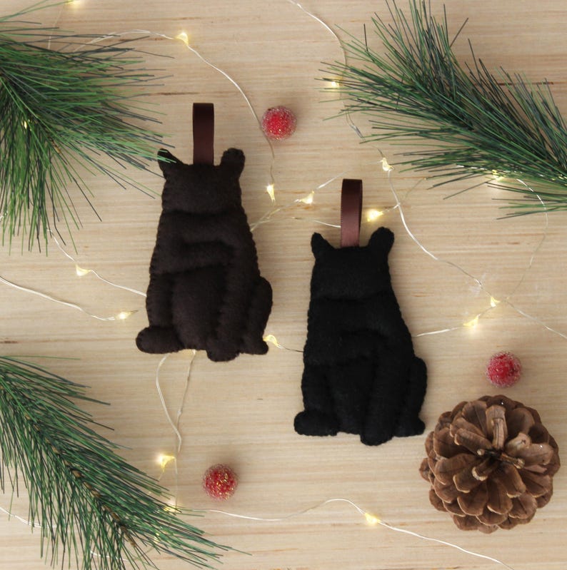 May include: Two felt bear ornaments, one brown and one black, hanging from brown ribbon. The bears are sitting with their paws tucked in front of them. The ornaments are on a wooden background with white lights and red berries.