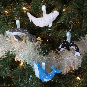 May include: Four felt whale ornaments hanging on a Christmas tree. The ornaments are a blue whale, a black and white orca, a gray humpback whale, and a white beluga whale. Each whale has a snowflake pattern.