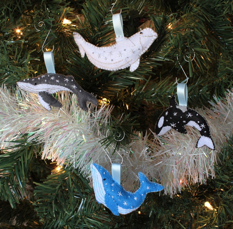 May include: Four felt whale ornaments hanging on a Christmas tree. The ornaments are a blue whale, a black and white orca, a gray humpback whale, and a white beluga whale. Each whale has a snowflake pattern.