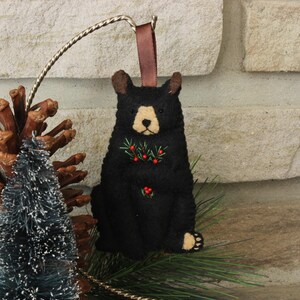 May include: Black felt ornament of a bear with red berries and green leaves embroidered on its chest. The bear is sitting upright with its paws visible. The ornament has a brown ribbon for hanging.