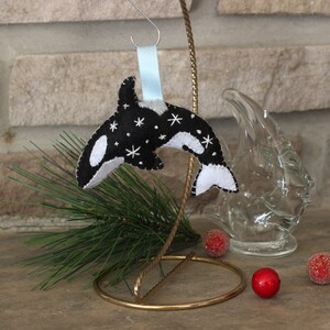 May include: Black and white felt ornament of an orca whale with white stars on a black background. The ornament is hanging from a blue ribbon on a gold metal stand.